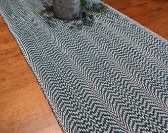 May include: A long, rectangular table runner featuring a woven chevron pattern in deep forest green and off-white threads. The fabric has a textured, dense weave. A galvanized metal pot containing a green leafy plant sits on the runner.
