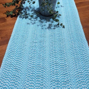 May include: A rectangular table runner featuring a woven pattern of light blue and white threads. The textile has a consistent textured surface throughout its length. A small metal pot containing a green leafy plant sits on the runner.
