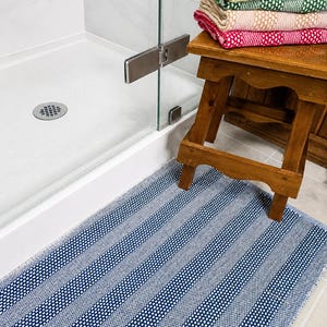 May include: A rectangular bath mat with a blue and white woven striped pattern lies on the floor. Beside it, a wooden stool holds a stack of folded towels in various colors including light blue, maroon, yellow, green, pink, and red.