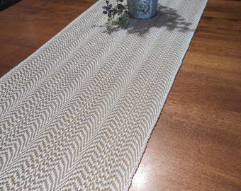 May include: A long, rectangular table runner featuring a woven chevron pattern in alternating shades of tan and white. The runner is laid flat, and a small, silver-colored metal bucket containing a green leafy plant sits on its surface.