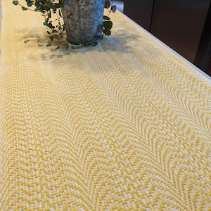 May include: A yellow and white woven table runner with a chevron pattern. The runner is on a table with a potted plant in the background.