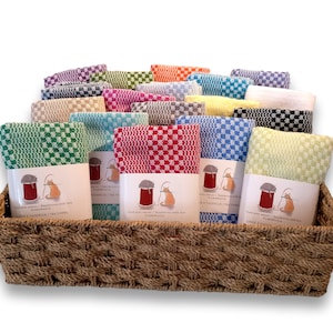 May include: A collection of folded checkered tea towels in various colors, including green, red, blue, and yellow, arranged in a woven brown basket. Each towel features a white paper band with an illustration of a cat and a spool of thread.