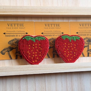 May include: Two heart-shaped beaded strawberry designs with red beads and green tops. The strawberries are displayed on a wooden shelf with business cards that read "VETTIE TULE RIVER YOKUT Handmade Art & Crafts."