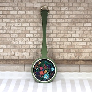May include: A long-handled olive green wooden paddle with a circular base featuring a dark blue center panel decorated with a vibrant, multi-colored floral pattern of red, orange, yellow, light blue, and green flowers and leaves, topped with a gold hook.