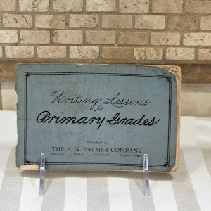 May include: A vintage blue rectangular book titled Writing Lessons for Primary Grades, published by The A. N. Palmer Company. The cover features black text, a decorative border, and shows signs of wear along the edges and corners of the spine.