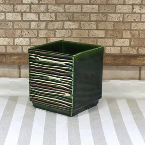 May include: A square-shaped ceramic planter with a deep forest green glaze. The front face features a textured pattern of horizontal, raised ridges in alternating shades of metallic copper, gold, and dark green, while the other sides are smooth.