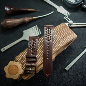 May include: Two brown leather watch straps with red stitching and a perforated design. The straps are on a wooden block with tools for leatherworking.