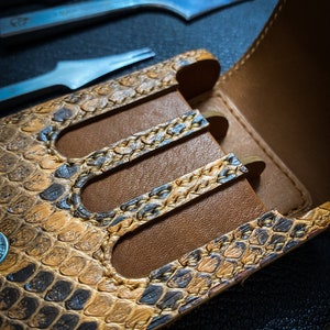 May include: A brown leather case with a snakeskin pattern and three slots for holding tools. The case is open and the slots are empty.