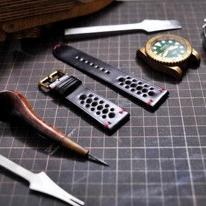 Puede incluir: Dos correas de reloj de cuero negro con costuras rojas y un patrón perforado. Las correas tienen hebillas doradas y están dispuestas sobre una superficie de rejilla negra.