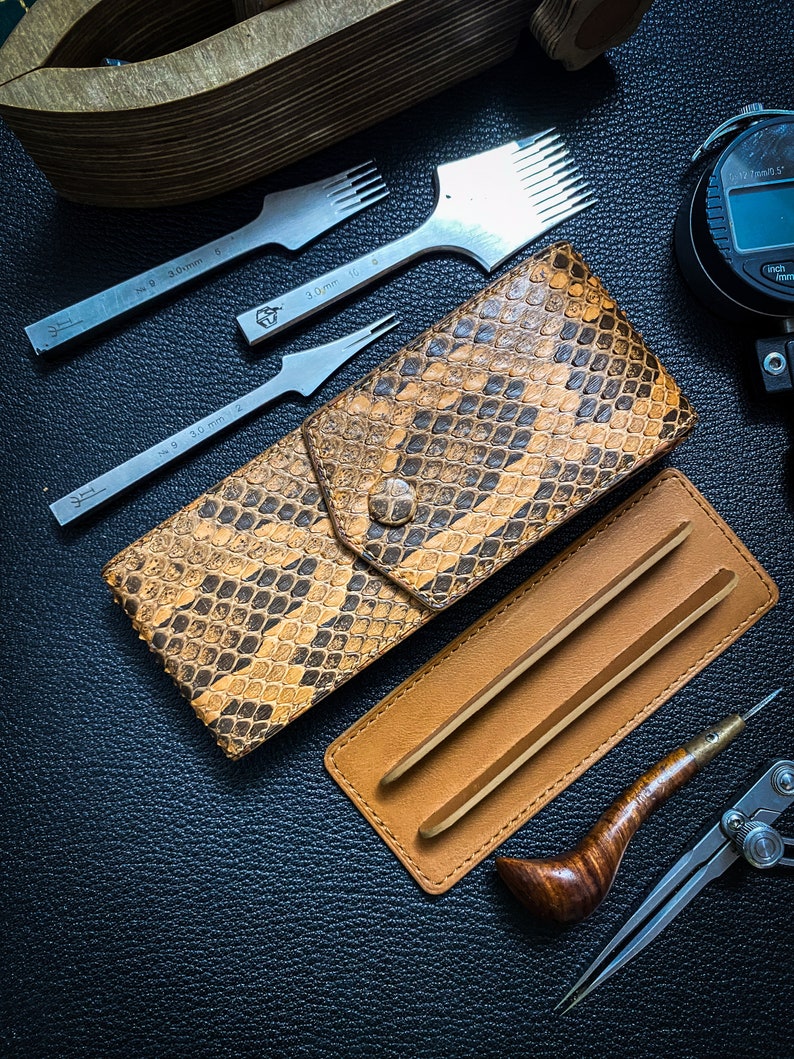 May include: A set of leatherworking tools including a leather punch, a stitching awl, a leather skiver, a pair of tweezers, a compass, and a leather wallet with a snakeskin pattern.