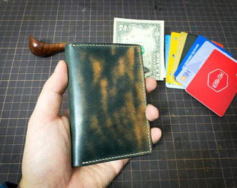May include: A green leather wallet with a distressed finish and white stitching. The wallet is being held in a hand.