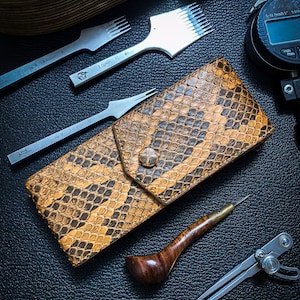 Op de afbeelding: Een bruine en beige slangenleren portemonnee met een klep sluiting. De portemonnee is omringd door leerbewerkingstools, waaronder een lederpons, een naald voor het stikken, een kompas en een leerdiktemeter.