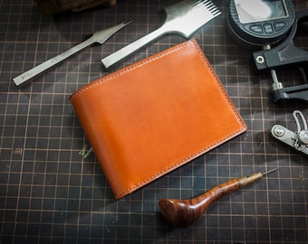 May include: A brown leather wallet with stitching around the edges. The wallet is lying on a black gridded surface with various leather working tools.
