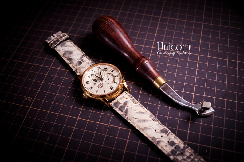 K&ouml;nnte beinhalten: Ein wei&szlig;es Lederarmband mit Krokodilmuster ist auf einer goldenen Uhr mit wei&szlig;em Zifferblatt zu sehen. Die Uhr hat r&ouml;mische Ziffern als Stundenmarkierungen und eine Mondphasenanzeige. Ein Lederbearbeitungswerkzeug ist ebenfalls auf dem Bild zu sehen.