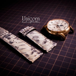 K&ouml;nnte beinhalten: Zwei wei&szlig;e Lederarmb&auml;nder mit einem strukturierten Muster. Die Armb&auml;nder liegen auf einem schwarzen Gitterhintergrund. Der Text "Unicorn handcraft leather" ist oben in der Mitte des Bildes sichtbar.