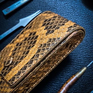 May include: A brown leather case with a snakeskin pattern. The case is closed and has a flap that folds over the top. The case is lying on a dark blue surface.