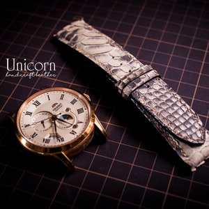 K&ouml;nnte beinhalten: Eine goldfarbene Armbanduhr mit wei&szlig;em Zifferblatt und r&ouml;mischen Ziffern als Stundenmarkierungen. Die Uhr verf&uuml;gt &uuml;ber eine Mondphasenanzeige und ein Datumsfenster. Die Uhr wird mit einem hellgrauen Armband aus Schlangenleder geliefert.