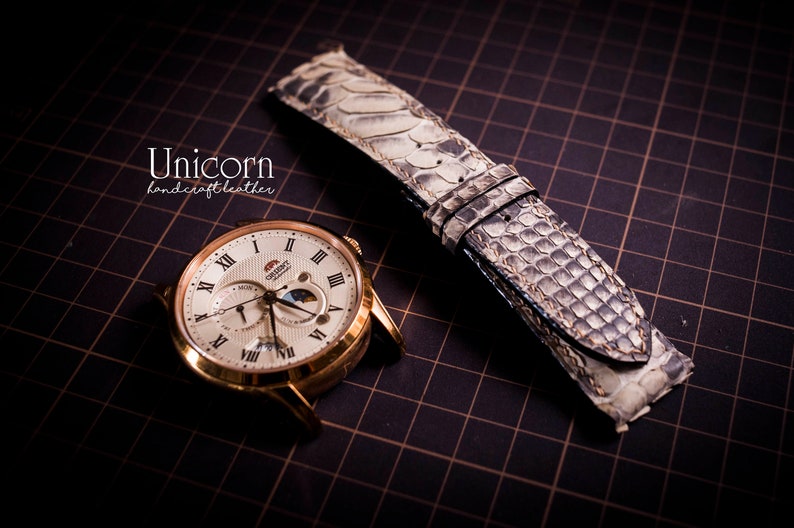 K&ouml;nnte beinhalten: Eine goldfarbene Armbanduhr mit wei&szlig;em Zifferblatt und r&ouml;mischen Ziffern als Stundenmarkierungen. Die Uhr verf&uuml;gt &uuml;ber eine Mondphasenanzeige und ein Datumsfenster. Die Uhr wird mit einem hellgrauen Armband aus Schlangenleder geliefert.