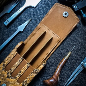 May include: A brown leather case with a snakeskin pattern, open to reveal three metal tools. The case has a snap closure and a metal button. The tools are for leatherworking and include a stitching awl, a stitching groover, and a stitching chisel.