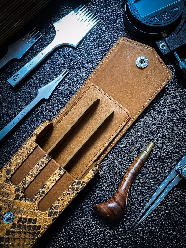 May include: A brown leather case with a snakeskin pattern, open to reveal three metal tools. The case has a snap closure and a metal button. The tools are for leatherworking and include a stitching awl, a stitching groover, and a stitching chisel.