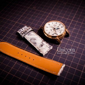 K&ouml;nnte beinhalten: Zwei Uhrenarmb&auml;nder, eines ist ein hellbraunes Lederarmband mit wei&szlig;en N&auml;hten und das andere ist ein wei&szlig;es Schlangenlederarmband. Die Armb&auml;nder befinden sich auf einem schwarzen Gitterhintergrund.