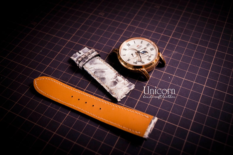 K&ouml;nnte beinhalten: Zwei Uhrenarmb&auml;nder, eines ist ein hellbraunes Lederarmband mit wei&szlig;en N&auml;hten und das andere ist ein wei&szlig;es Schlangenlederarmband. Die Armb&auml;nder befinden sich auf einem schwarzen Gitterhintergrund.