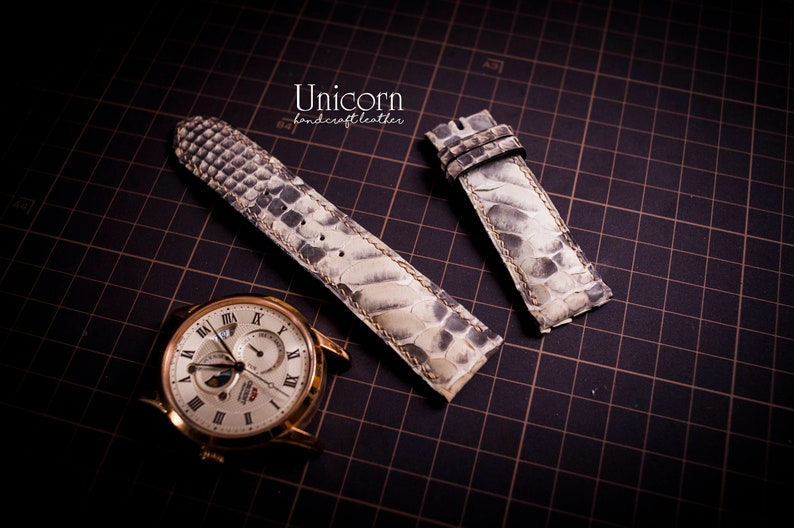 K&ouml;nnte beinhalten: Zwei wei&szlig;e Lederarmb&auml;nder mit Schlangenhautmuster. Die Armb&auml;nder liegen auf einem schwarzen Gitterhintergrund. Der Text "Unicorn handcraft leather" ist oben in der Mitte des Bildes sichtbar.