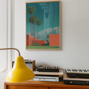 May include: A framed art print featuring a desert landscape with palm trees, red buildings, and mountains under a teal sky. The print is in a wooden frame. A yellow lamp, audio equipment, and a keyboard are on a wooden cabinet.
