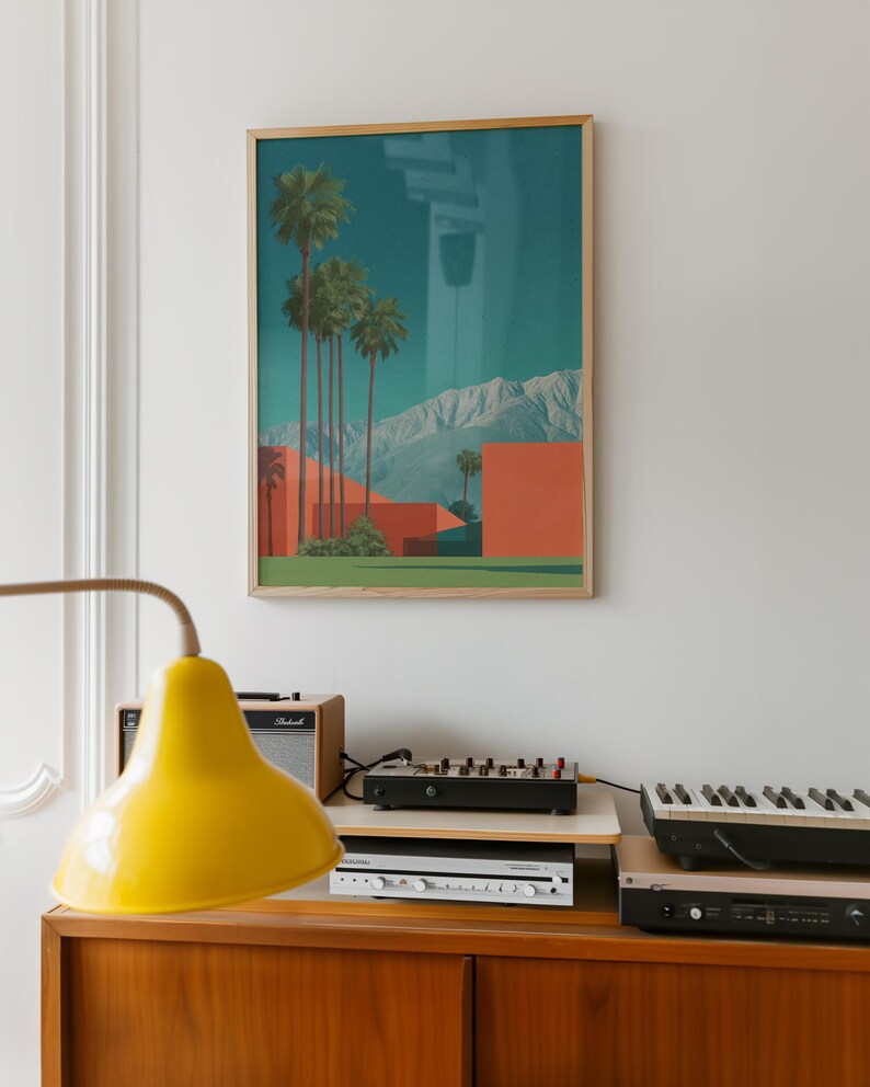 May include: A framed art print featuring a desert landscape with palm trees, red buildings, and mountains under a teal sky. The print is in a wooden frame. A yellow lamp, audio equipment, and a keyboard are on a wooden cabinet.