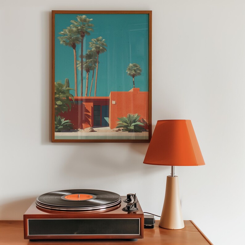 May include: A framed print of a mid-century modern building with palm trees and a turquoise sky hangs above a record player and a lamp with an orange shade. The record player is brown with a black record on top. The lamp has a beige base.