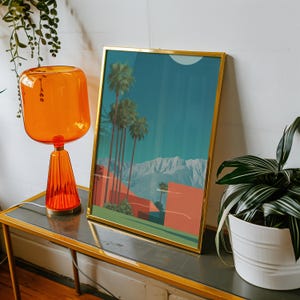 May include: A framed art print with a gold frame depicts a desert landscape with palm trees, mountains, and buildings in shades of orange and teal. An orange glass lamp and a potted plant are also visible.