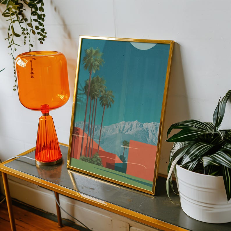 May include: A framed art print with a gold frame depicts a desert landscape with palm trees, mountains, and buildings in shades of orange and teal. An orange glass lamp and a potted plant are also visible.