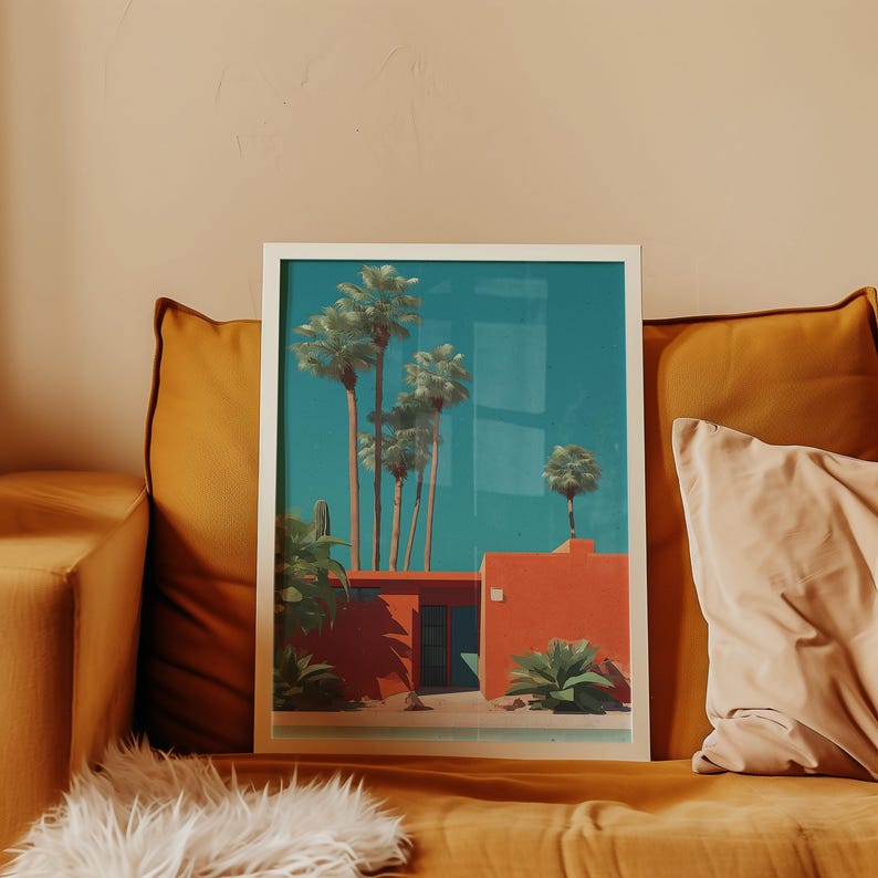 May include: A framed art print featuring a desert scene with palm trees and a red building against a teal sky. The artwork is displayed on a mustard-colored couch with a white frame.