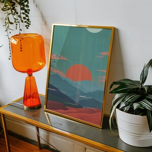 May include: A framed art print with a gold frame depicts a landscape scene with a large orange sun setting over blue mountains. The print is displayed on a gold-trimmed table next to an orange glass lamp and a potted plant.