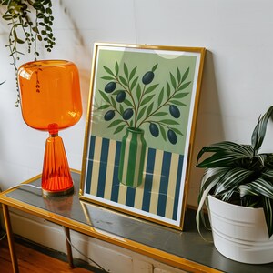 May include: A framed art print featuring a green vase with olive branches and olives. The background has a striped pattern in blue and yellow. The print is in a gold frame and is displayed on a table with an orange lamp.