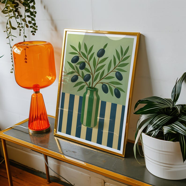 May include: A framed art print featuring a green vase with olive branches and olives. The background has a striped pattern in blue and yellow. The print is in a gold frame and is displayed on a table with an orange lamp.