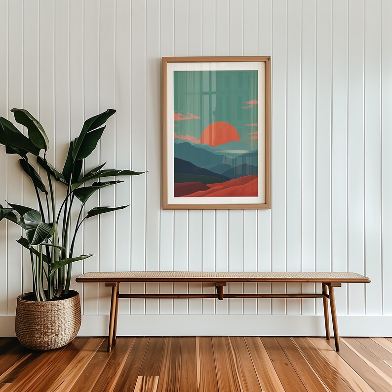 May include: A framed landscape art print featuring a stylized sunset over mountains. The artwork uses a palette of orange, teal, and red hues. The print is displayed on a white wall above a wooden bench with a woven seat.