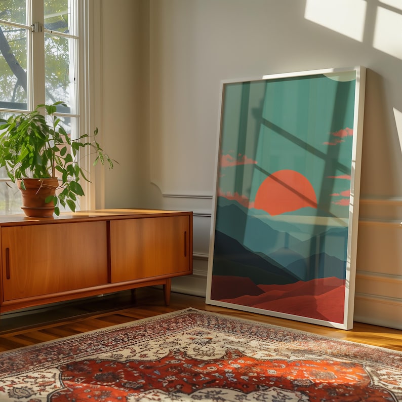 May include: A framed art print featuring a sunset over a mountain range. The artwork has a color palette of orange, teal, and red. The print is leaning against a wall next to a wooden cabinet and a patterned rug.