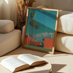 May include: A framed art print featuring a desert landscape with palm trees, red buildings, and mountains under a teal sky. The artwork is in a wooden frame and rests on a beige surface near an open book.