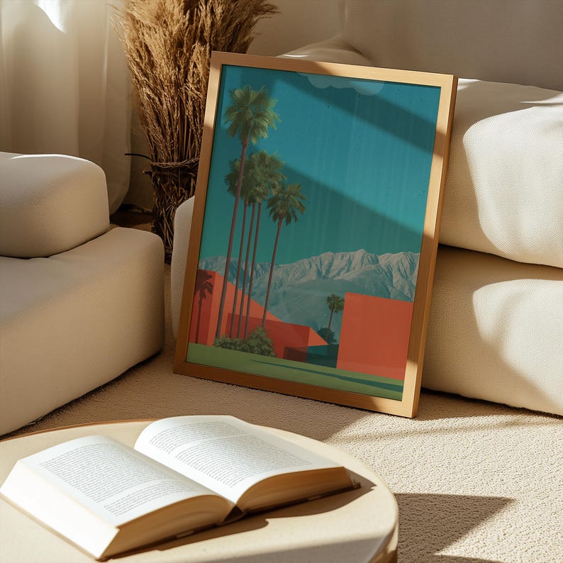 May include: A framed art print featuring a desert landscape with palm trees, red buildings, and mountains under a teal sky. The artwork is in a wooden frame and rests on a beige surface near an open book.