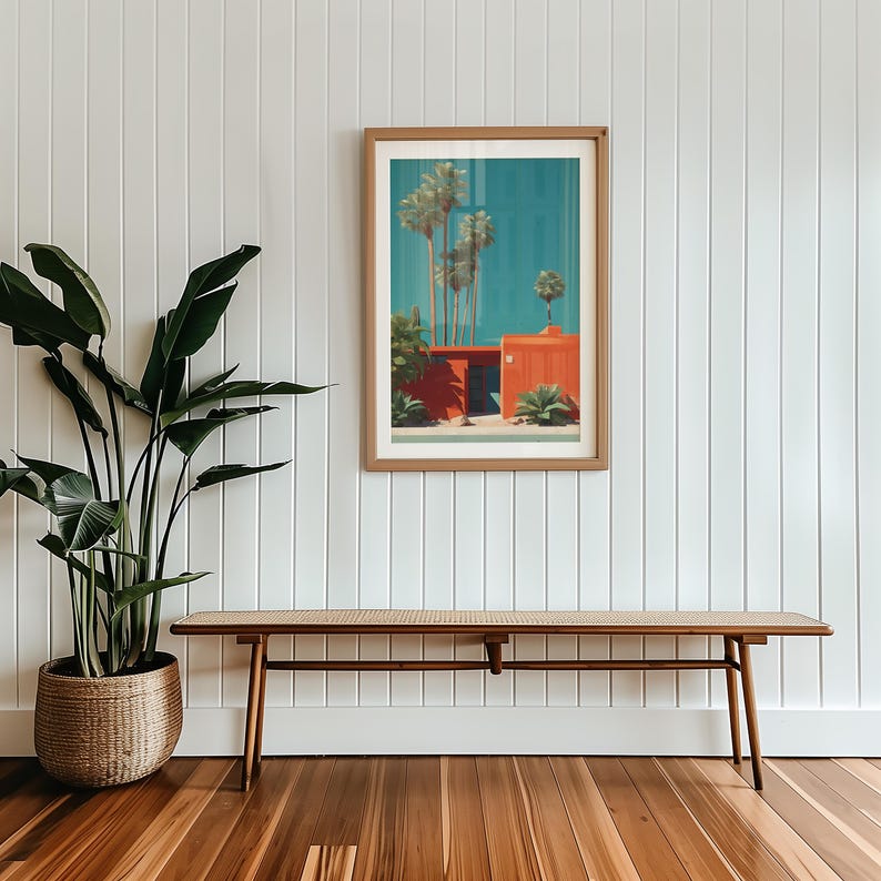May include: A framed art print depicting a mid-century modern style building with palm trees against a teal sky. The artwork is in a wooden frame, hanging on a white paneled wall. A wooden bench with a woven seat sits below the artwork.