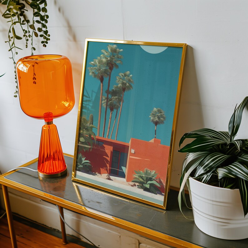 May include: A framed art print with a gold frame depicts a desert scene with palm trees and a red building against a blue sky. An orange glass lamp and a potted plant sit on a table.
