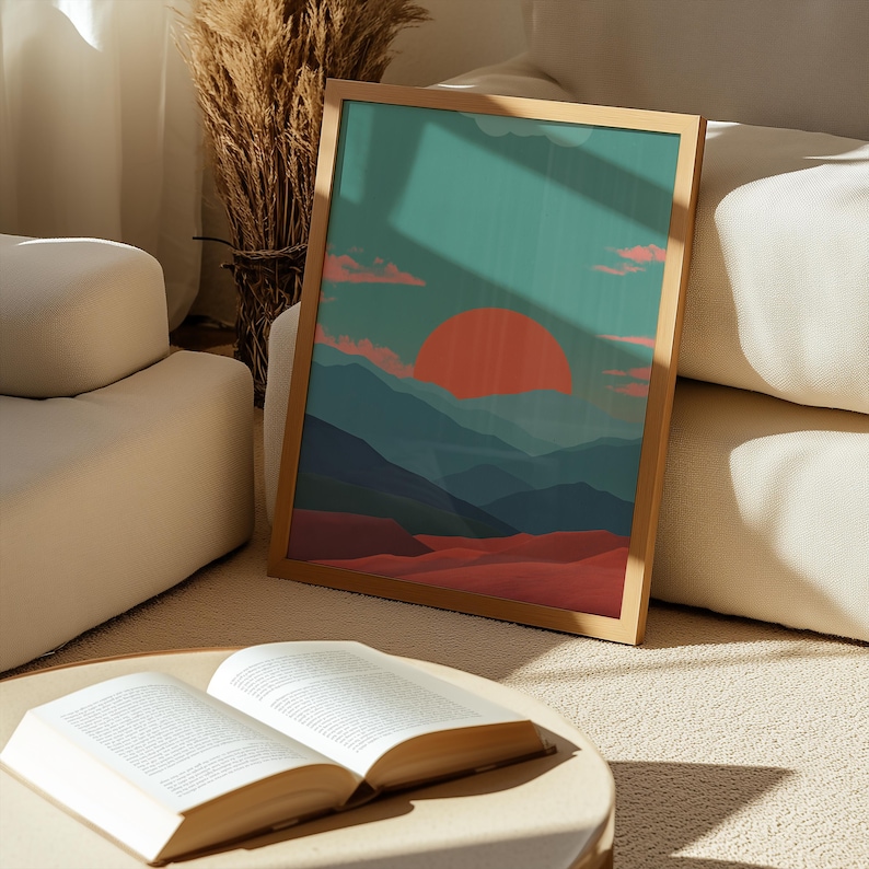 May include: A framed landscape art print featuring a sunset over mountains. The artwork has a teal sky, a large orange sun, and layers of blue mountains. The foreground shows red dunes. The frame is a light brown color.