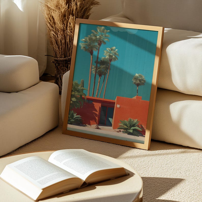 May include: A framed art print featuring a mid-century modern style illustration of palm trees and a red building against a teal sky. The artwork is in a wooden frame and rests on a beige surface near an open book.
