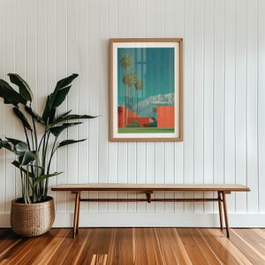 May include: A framed art print featuring a desert landscape with palm trees, red buildings, and mountains under a blue sky. The print is displayed above a wooden bench with a woven seat, next to a large potted plant.