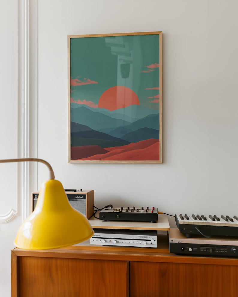 May include: A framed art print depicting a sunset over a mountain range. The artwork features a vibrant orange sun, layered blue mountains, and reddish-brown foreground. The frame is a light wood color. The print hangs above a wooden cabinet with electronic equipment.