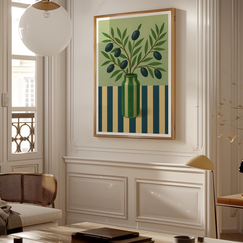 May include: A framed art print featuring a green vase with olive branches and black olives. The vase sits on a striped background of blue and yellow. The artwork is displayed in a room with white walls and a gold frame.