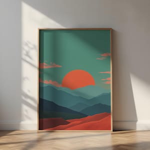 May include: A framed art print featuring a stylized landscape. The artwork depicts a large orange sun setting over layered blue mountains and red-orange dunes, set against a teal sky with pink clouds. The frame is a light brown color.