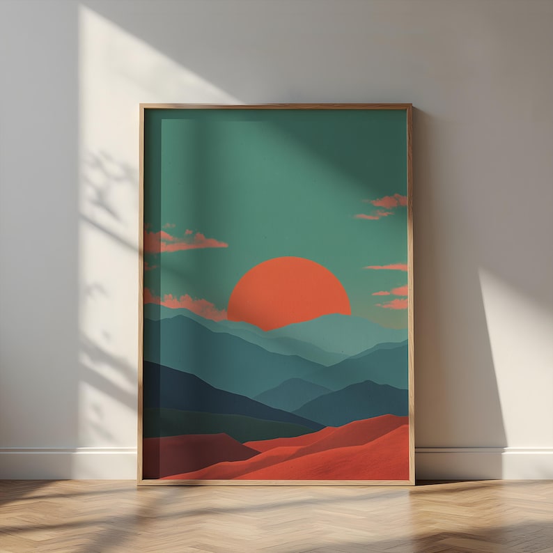 May include: A framed art print featuring a stylized landscape. The artwork depicts a large orange sun setting over layered blue mountains and red-orange dunes, set against a teal sky with pink clouds. The frame is a light brown color.