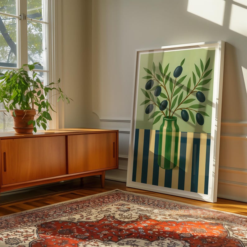 May include: A framed art print featuring a green vase with olive branches and black olives. The background is a light green with a striped pattern in blue and yellow. The artwork is leaning against a white wall.